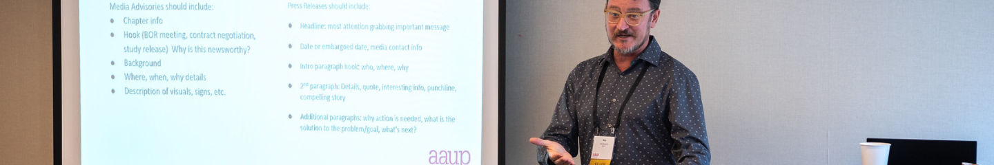 Presenter wearing a name badge and gesturing with both hands stands between podium on the right and screen with a slide presentation with information about press releases on the left