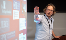 Presenter stands to the right of a screen with logos of conservative donors and holds his hand out toward the screen