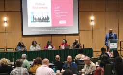 Lecture hall with crowd facing a table of panelists next to a presenter at a podium; slide show lists bullet points headed "Political Environment"