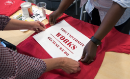 On a table, there is a silkscreen template that says "THIS UNIVERSITY WORKS BECAUSE WE DO" is placed on a red T-shirt by two people whose arms are visible as they adjust the template from either side of the table