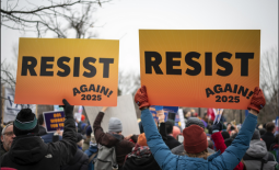 a crowd at a winter demonstration shows two people from the back holding up signs with an orange and yellow gradient that say RESIST in the middle and AGAIN 2025 in the lower right