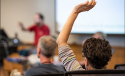Rear view of man raising his arm in a lecture hall, facing a presenter and a screen with a slide presentation