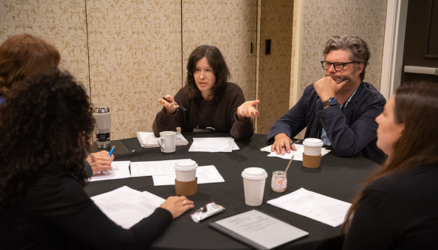 Five people sit around a table in a conference room with notepads and coffee cups on the table. A woman talking gestures with her hands and a man sitting next to her has his chin in his hand while others face them.