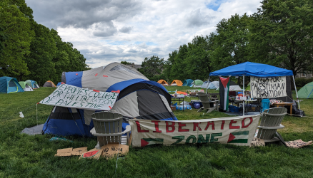Encampment on a campus lawn shows tents creating an enclosure with a banners declaring "Liberated Zone" and "People's College for the Liberation of Palestine"