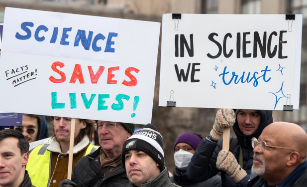winter scene of protest with signs affirming the value of trust, including "Science Saves Lives!" and "In Science We Trust"